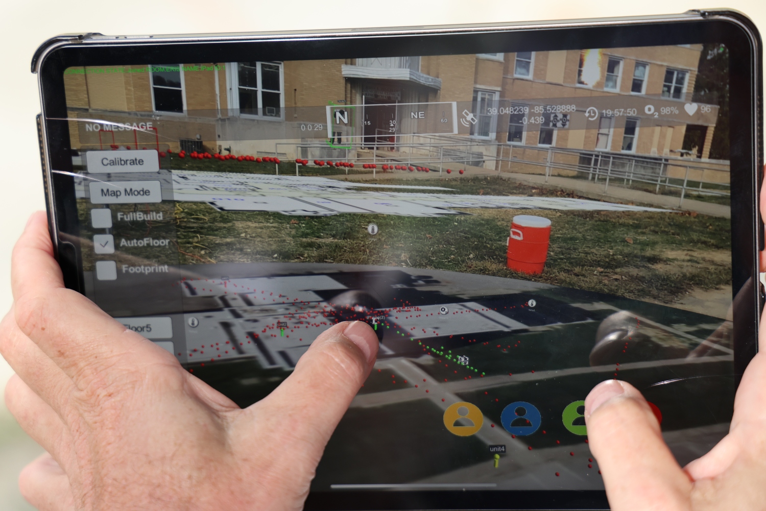 A person holds a tablet that shows both the outside structure of a brick building as well as the aerial view. Red and green location tracking dots are scattered throughout the view.