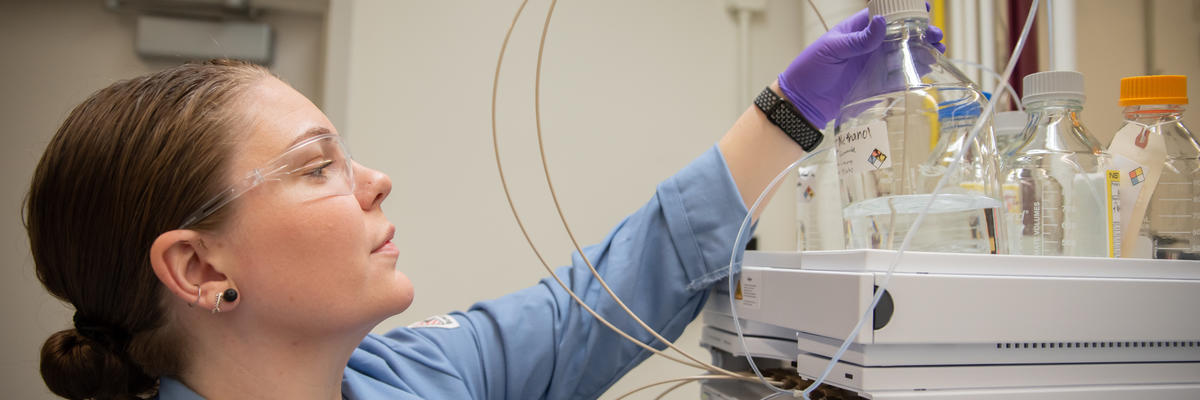 阿利克斯·罗德瓦（Alix Rodowa）在实验室里戴着安全眼镜和手套，伸手去拿放在一件科学设备上的透明液体容器。 