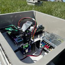 An open gray box filled with circuitry, wires, sensors and a single-board computer sits outside on a lawn.