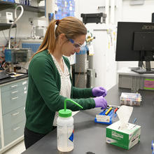 Briana Capistran wears safety glasses and purple gloves as she prepares small vials at a lab table. 