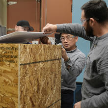 Three men wearing safety glasses stand around a wooden box they are assembling in the lab.