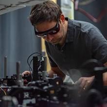 Krister Shalm wears safety glasses as he leans over a lab table covered with electronics equipment.