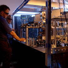 Two researchers in safety glasses stand at the edge of a metal table holding electronic and laser devices, wiring and metal structures.