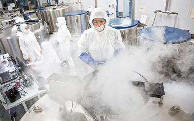 Giant metal tubs are being opened and white vapor is puffing up into the face of one researcher who is in a protective suit with hood, gloves and eye protection. In the background, several other suited researchers discuss something.