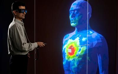 A researcher in safety glasses stands holding a remote control near a large blue model of a human head and torso with red marks on the chest. 
