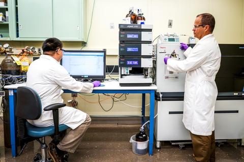 NIST research chemists Yuxue Liang (left) and Yamil Simon (right) in white lab coats acquiring data from a mass spectrometer.