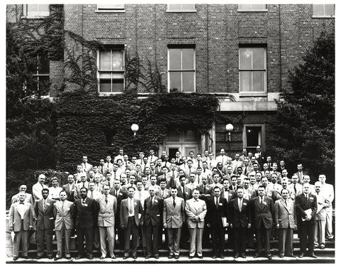 Men pose outside a brick building in a historical black-and-white photo. 
