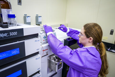 A woman in a purple coverall reaches up to work on scientific equipment.