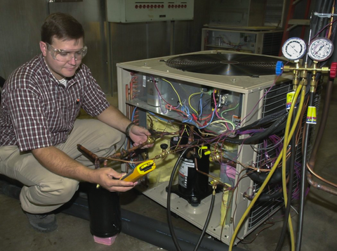 A man in safety goggles crouches at the back of an open air-conditioning unit, looking at a handheld tester.