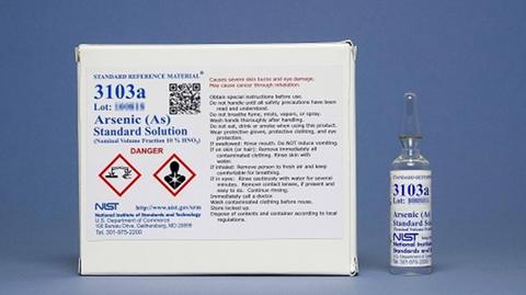 Photograph of SRM 3103a showing a labeled clear glass ampoule and labeled box against a blue background.