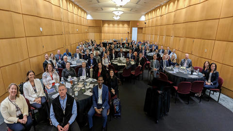 Group picture of attendees sitting in round tables smiling at the 2023 Manufacturing USA Network Meeting 