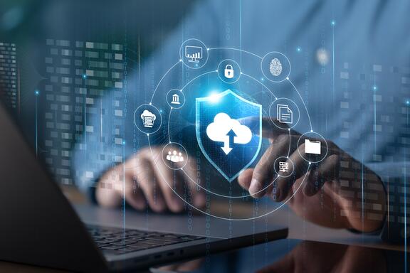 Illustration shows cloud, cybersecurity and identity icons floating over an open laptop with a person's hands at the keyboard.