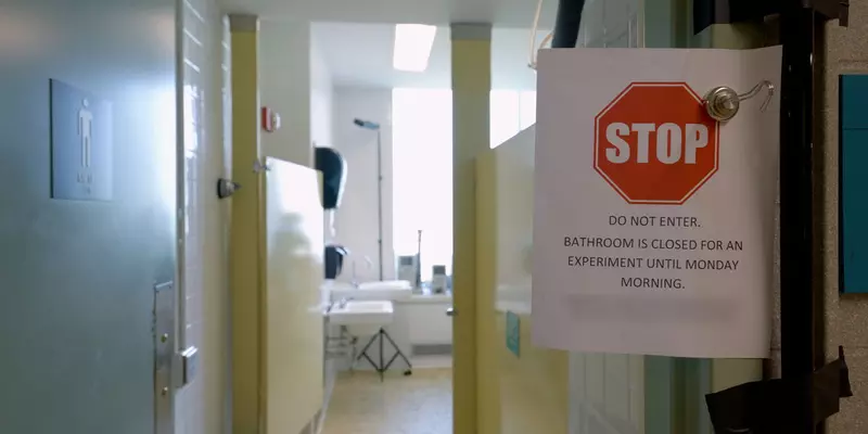 公共厕所的一扇敞开的门前夹着一个纸牌。告示牌上写着：“站住，请勿入内。浴室在周一早上之前都不开放进行实验。”