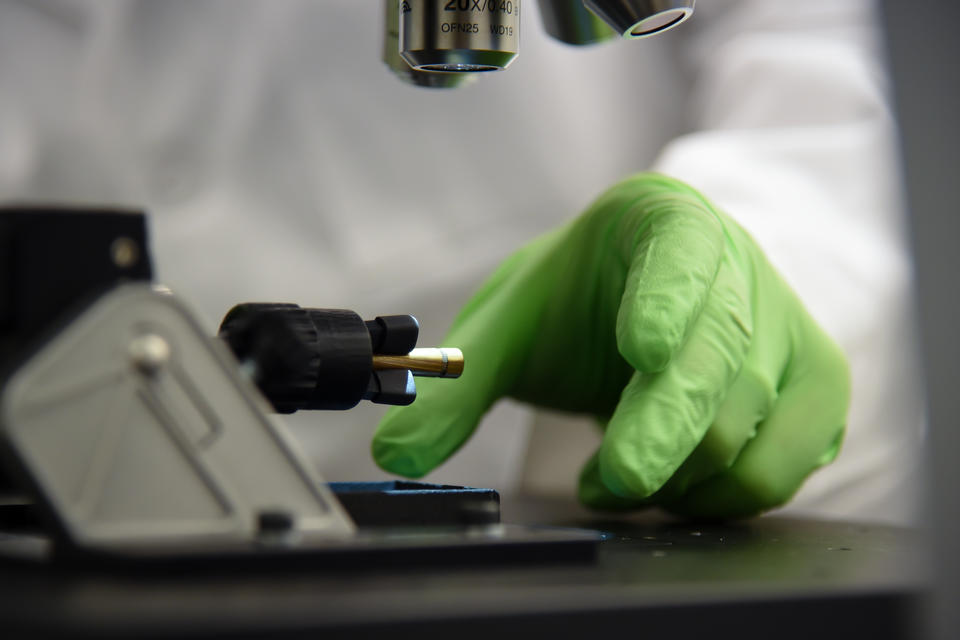 Person wearing green glove reaches under microscope to remove sample.