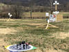 An aerial drone sits on a launch pad before lifting off to start the NIST performance test course. Three sections of the course, poles with white bucket-shaped targets attached, are seen in the background.