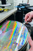 A circular silicon wafer with rainbow reflections lies flat while a worker uses a small tool to touch the edge. 