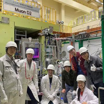 Seven researchers wearing white hard hats and lab coats pose for a group photo in front of large pieces of lab equipment. Sign above says: Muon D2.