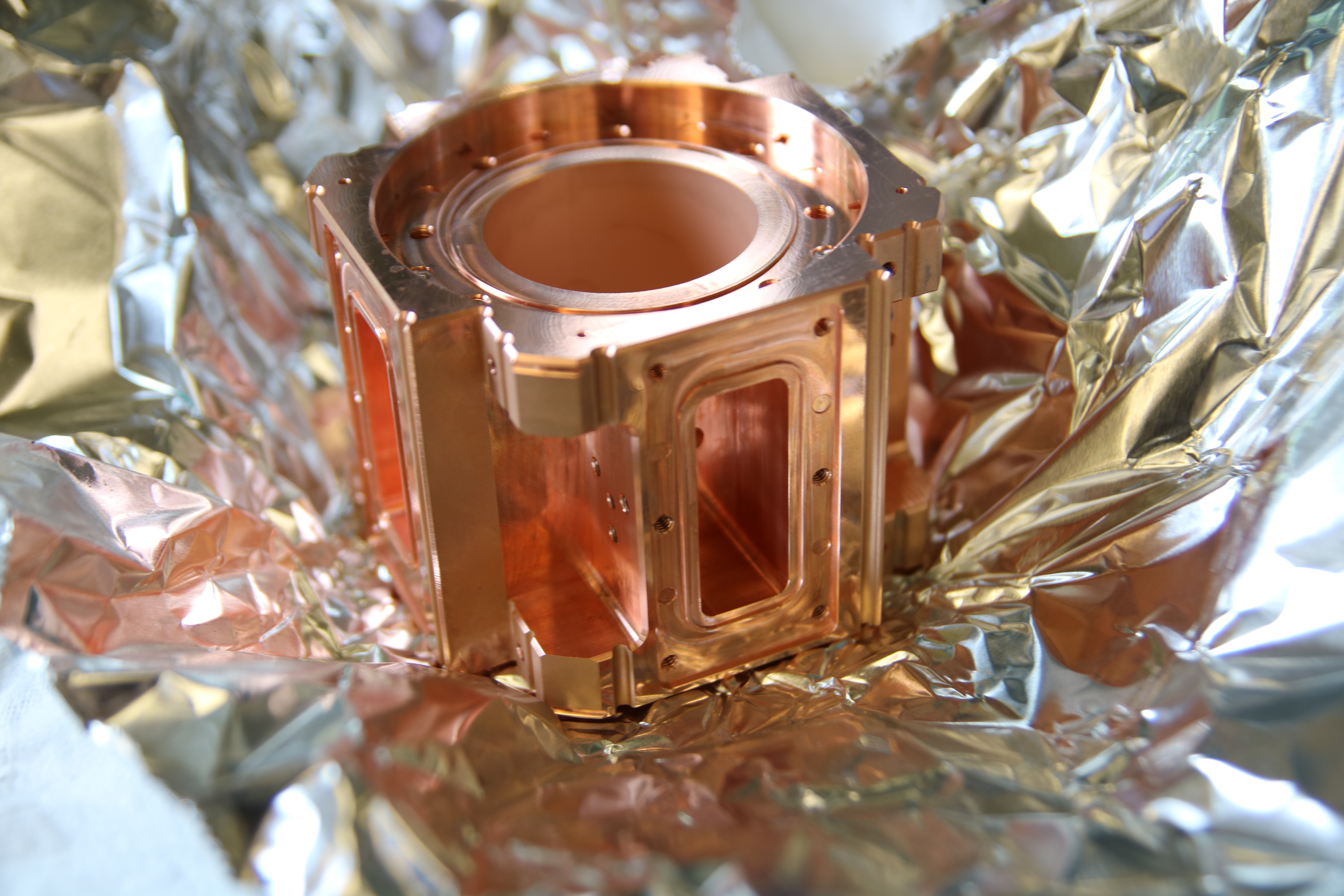 Approximately cube-shaped copper-colored metal object with circular opening on the top sits on a piece of aluminum foil.