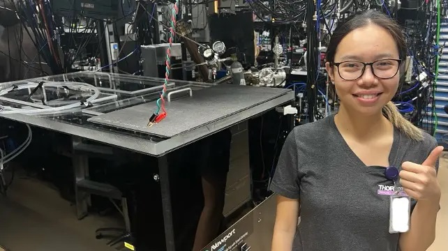 A woman poses in a lab full of electronics and wiring, making a thumbs-up gesture. 