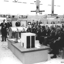 Two men stand in front of poster boards. A bunch of men are seated watching them present.