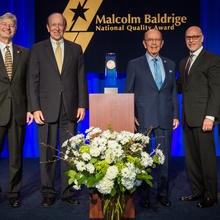 Center: Baldrige Award on a podium. Left: 2 men in suits. Right: 3 men in suites