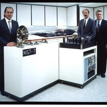 Three men stand behind a glass tube with equipment inside