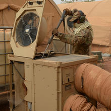 A man in brown camouflage clothing, including baseball cap, gloves and ear protection, uses a long pole to clean the inside of an HVAC unit outside a military tent.