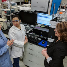 Carolyn Burdette, Laura Regalado and Katrice Lippa having an animated discussion in the lab. Behind them is a computer screen and other laboratory equipment, including a liquid chromatography mass spectrometer. 