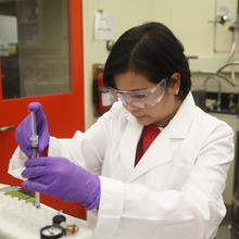 A woman with goggles prepares a sample near a large instrument.