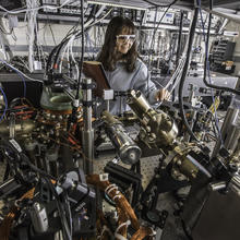 woman in goggles standing behind a lab table filled with equipment and wires. She has a lab notebook in her hand.