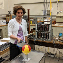 Woman standing in a lab holds a rectangular radiation detector near a metal globe that has been painted with a radiation symbol