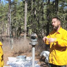 NIST engineer in yellow jacket stands to right of BOB glass globe. To his right and left are two men.