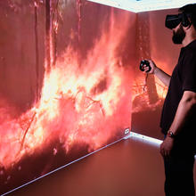 A profile of a man wearing VR equipment is seen against two large screens displaying the view from within a forest fire.