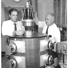 Two men stand on either side of a large cylindrical device.