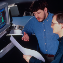 Two men look at a printout in front of a computer screen