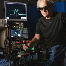 Man in black shirt and dark glasses facing camera is adjusting a mirror on a table of lasers and optics. On the left behind him are several electronics boxes topped by a computer monitor showing a blue signal that looks like cats ears.
