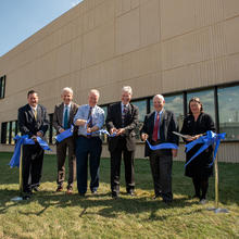 Six people stand in front of a tan building, cutting a blue ribbon