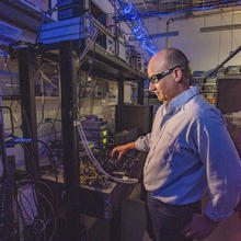 A man in safety glasses stands near a rack of equipment in a darkened lab.