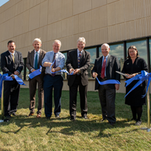 Six people stand in front of a large tan building, cutting a blue ribbon.