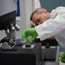 A man in a lab coat and green gloves places a bullet under a microscope.