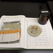 Several cigarettes, two extinguished butts, and a brown jar filled with cigarette smoke are sitting on top of a lab notebook full of notes. 