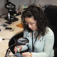A woman uses a soldering iron to attach wires to a circuit board.