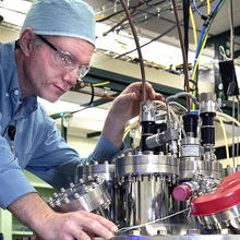 man with goggles, blue shirt and blue cap adjusts a pipe on top of a gleaming aluminum ultra-high vacuum (UHV) metal deposition chamber