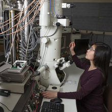 NIST researcher in a laboratory room looking at tall vertical microscope with multicolored wires emanating from it.