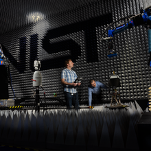 Researchers in large anechoic chamber with NIST logo on wall looking at robotic arms and equipment.