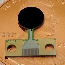 A small flat device with a round black part and a copper-colored rectangular part lies on top of a penny.