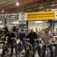 group of men parked on their bikes and smiling for the camera in a large guidehall. The sign hanging from the ceiling behind them reads "NIST NSLS-II Partner Beamlines, Bright light for material measurement"