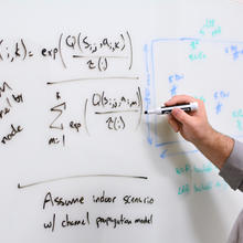 Man with dark hair writing mathematical formulas on a whiteboard on a wall.