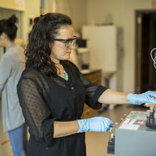 A woman with safety goggles and gloves stands at a lab bench.