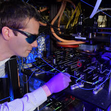 Stephen Eckel wears dark safety glasses as he peers into the complex CAVS apparatus in a darkened lab. 